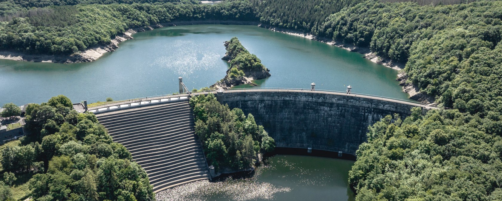 Luftaufnahme der Urfttalsperre mit der Urftstaumauer, umgeben von dicht bewaldeten Hügeln und blauem Wasser unter einem klaren Himmel., © Eifel Tourismus GmbH, Dennis Stratmann - finanziert durch REACT-EU