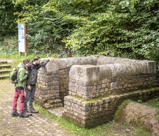 Zwei Personen stehen an einem historischen Brunnen im Grünen bei Nettersheim. Sie betrachten die Steinstruktur, umgeben von Bäumen und einer Infotafel., © Eifel Tourismus GmbH, Dominik Ketz